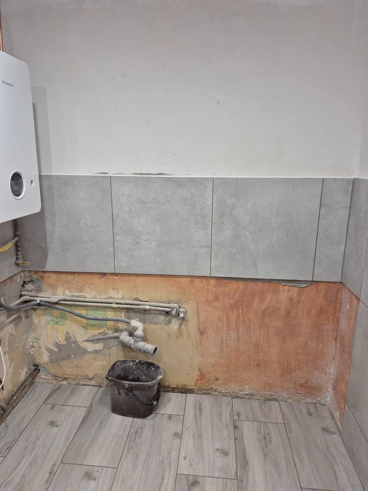 Remont łazienki w trakcie: ułożone szare płytki podłogowe imitujące drewno i częściowo położone płytki ścienne. Widoczne rury i wiadro z zaprawą.