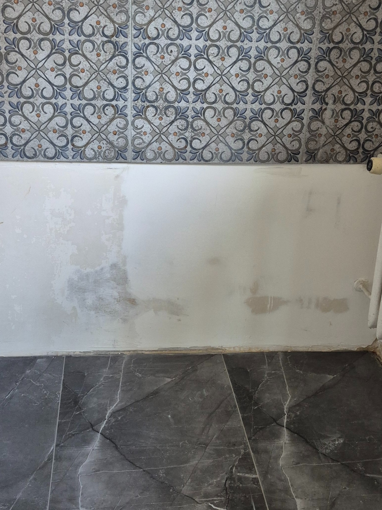 Ściana łazienki w trakcie przygotowania do położenia płytek, widoczne fragmenty szpachli i dekoracyjne płytki na górze. Na dole ciemna podłoga z płytek imitujących marmur.