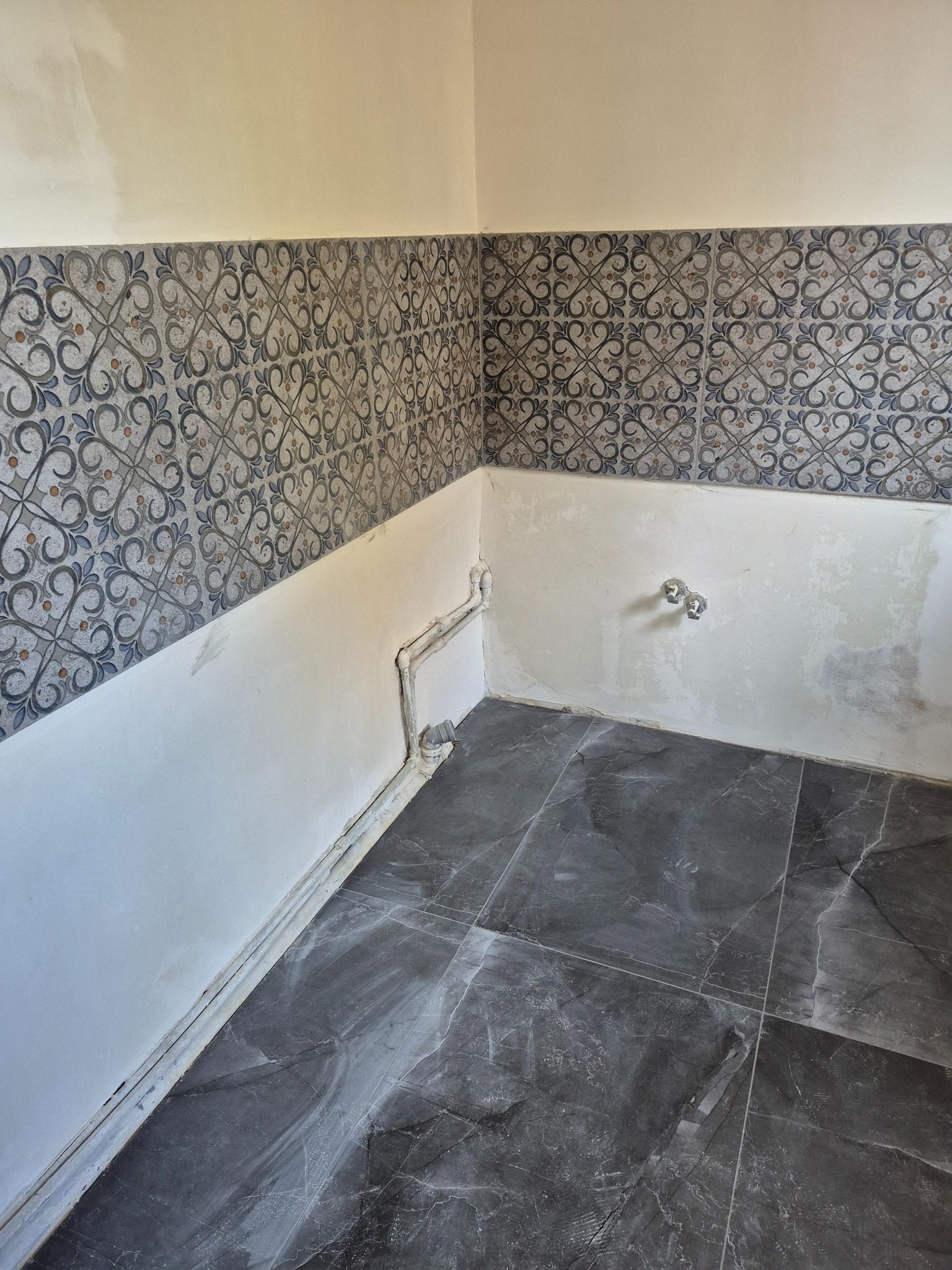 Łazienka w trakcie remontu: ułożone ciemne płytki podłogowe, częściowo położone dekoracyjne płytki ścienne, widoczne rury i przygotowane ściany do dalszych prac.