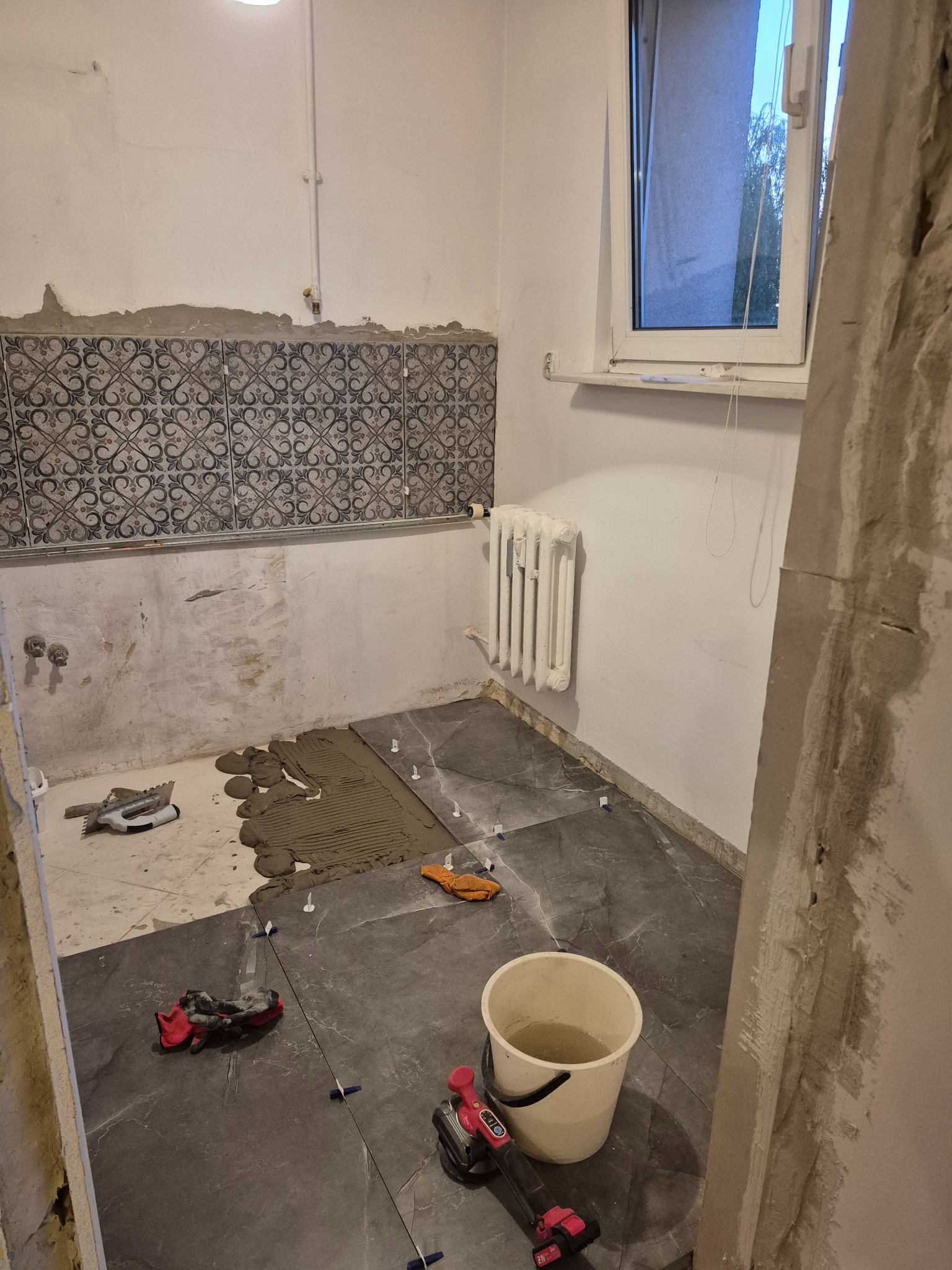 Łazienka w trakcie układania płytek podłogowych w odcieniach szarości. Widoczne narzędzia glazurnicze, wiadro z wodą i częściowo ułożone płytki na ścianie.