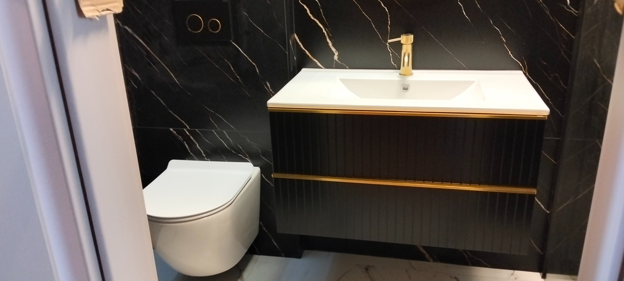Elegancka łazienka z czarnymi płytkami imitującymi marmur, białą ceramiką i złotymi akcentami na szafce umywalkowej i baterii. Nowoczesny design.