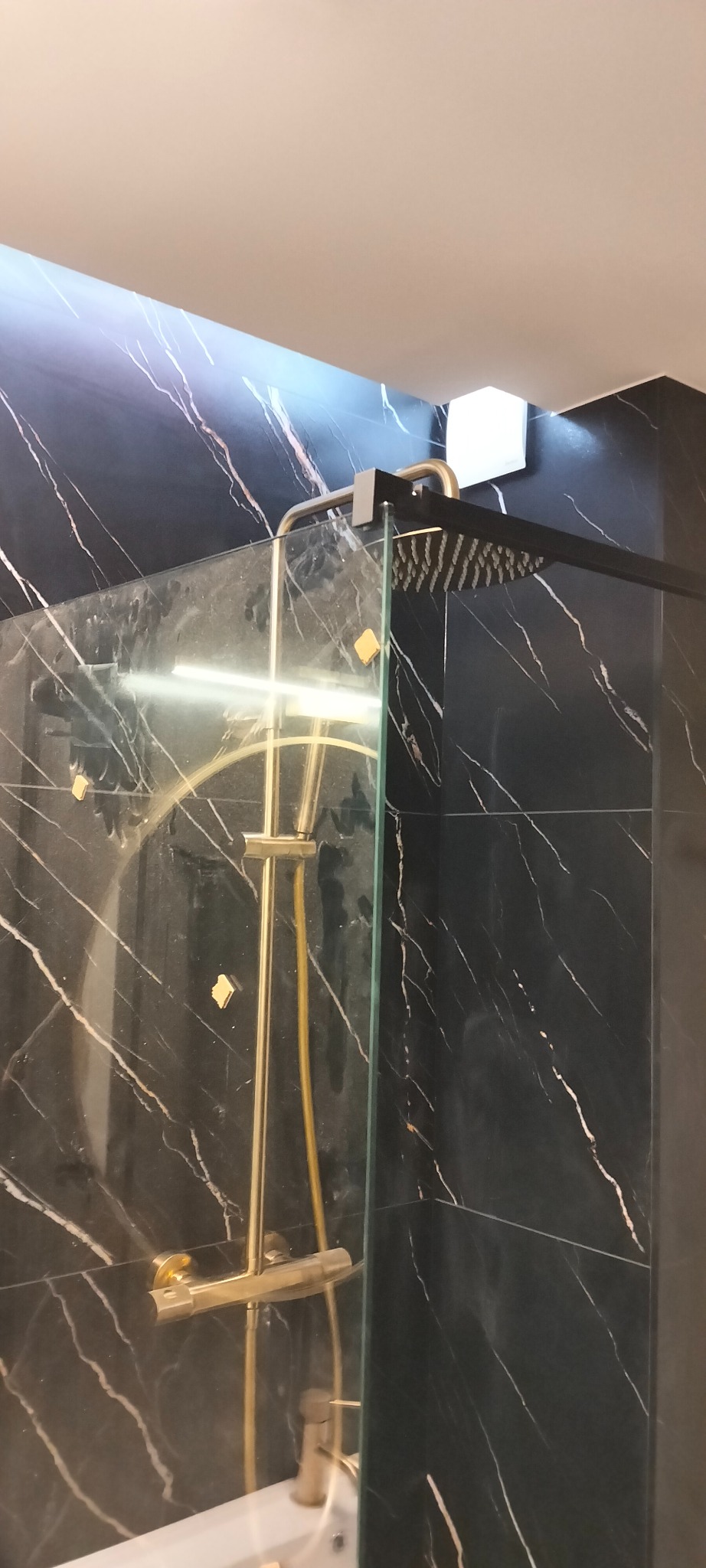 Nowoczesna kabina prysznicowa z czarnymi płytkami imitującymi marmur i złotą armaturą. Widoczny deszczownica i bateria. Minimalistyczny design.