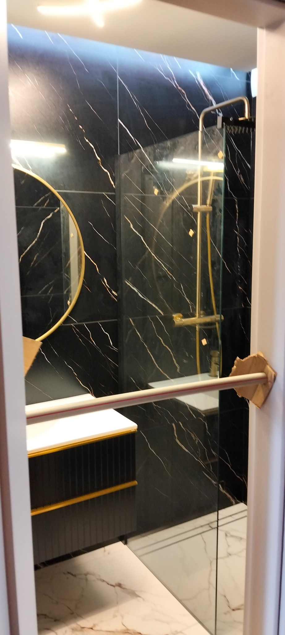 Wykończona łazienka z czarnymi płytkami imitującymi marmur, złotą armaturą i okrągłym lustrem. Widoczny fragment szafki pod umywalką i kabina prysznicowa.