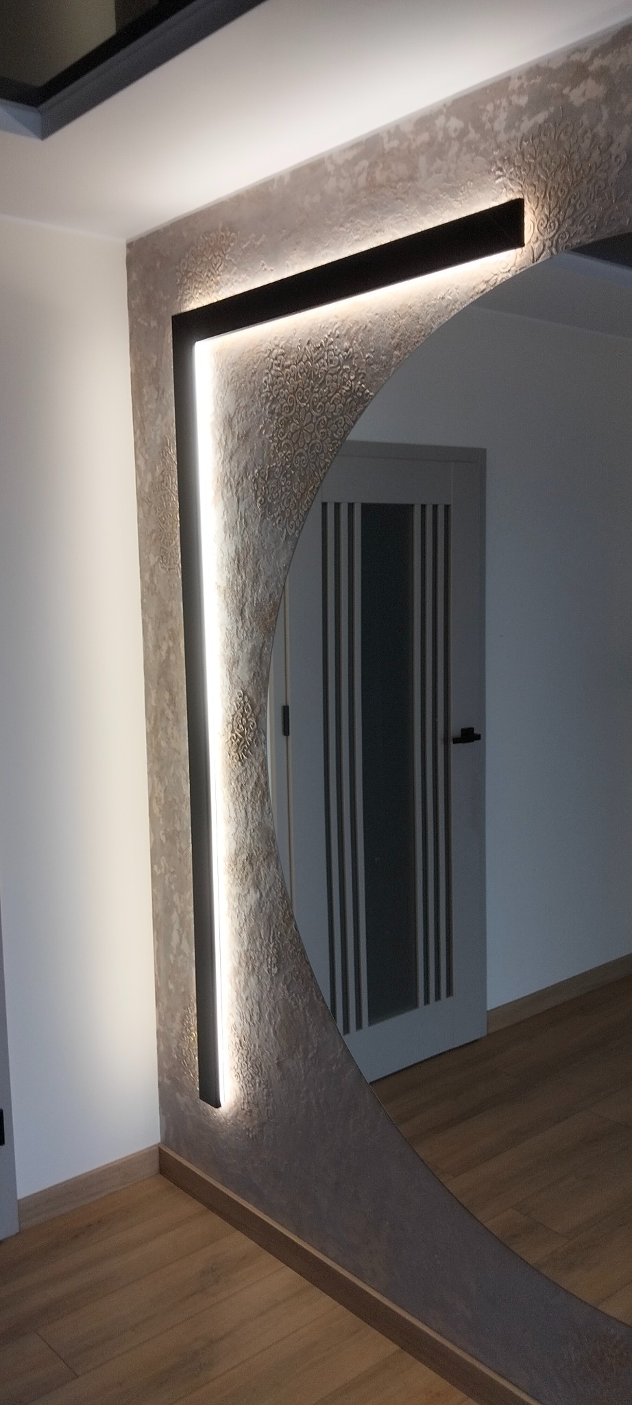 Oświetlone lustro z dekoracyjną ramą i wzorem na ścianie, odbijające drzwi w nowoczesnym wnętrzu. Ciepłe światło podkreśla fakturę ściany i dodaje elegancji.