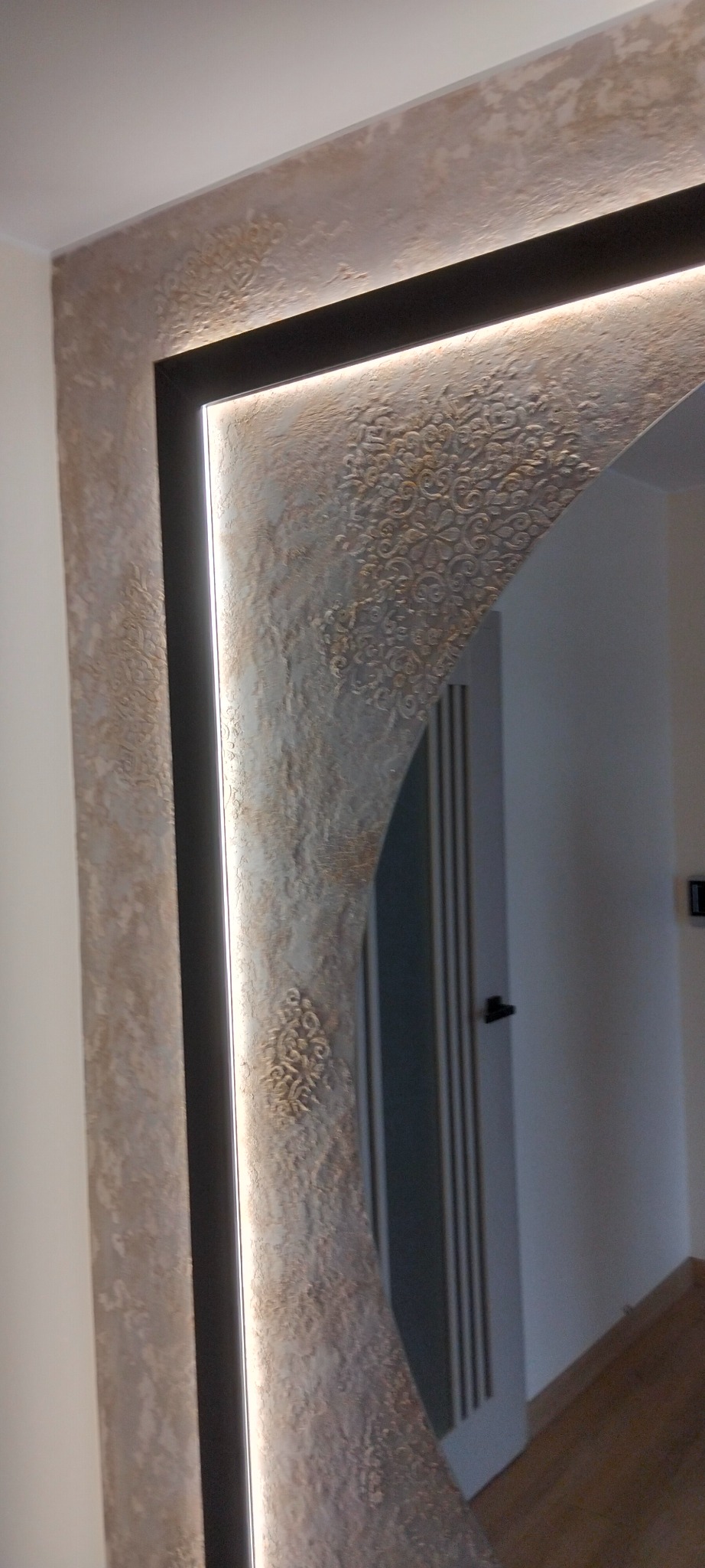 Dekoracyjna ściana z efektem stiuku w odcieniach beżu i brązu, oświetlona listwą LED w czarnej ramie, z widocznym fragmentem drzwi w tle.