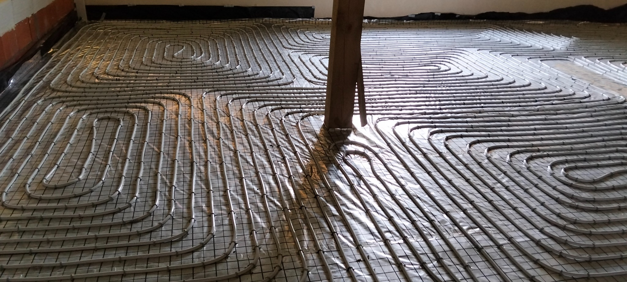 Ułożone rury ogrzewania podłogowego na folii, widoczne mocowania do siatki. W tle ściana i fragment sufitu. Centralnie pionowy słup konstrukcyjny.