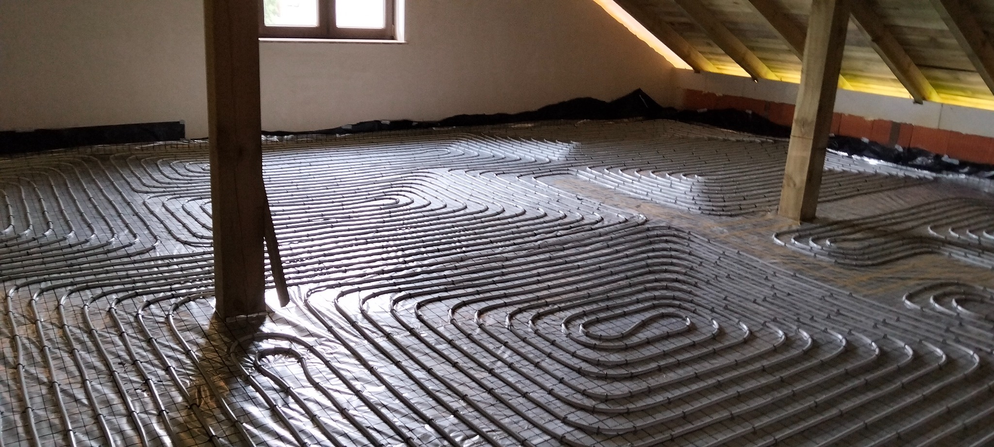 Instalacja ogrzewania podłogowego z rur na siatce, widok z góry, drewniany słup w kadrze, poddasze ze skosami, okno w tle. Etap prac przed wylaniem posadzki.