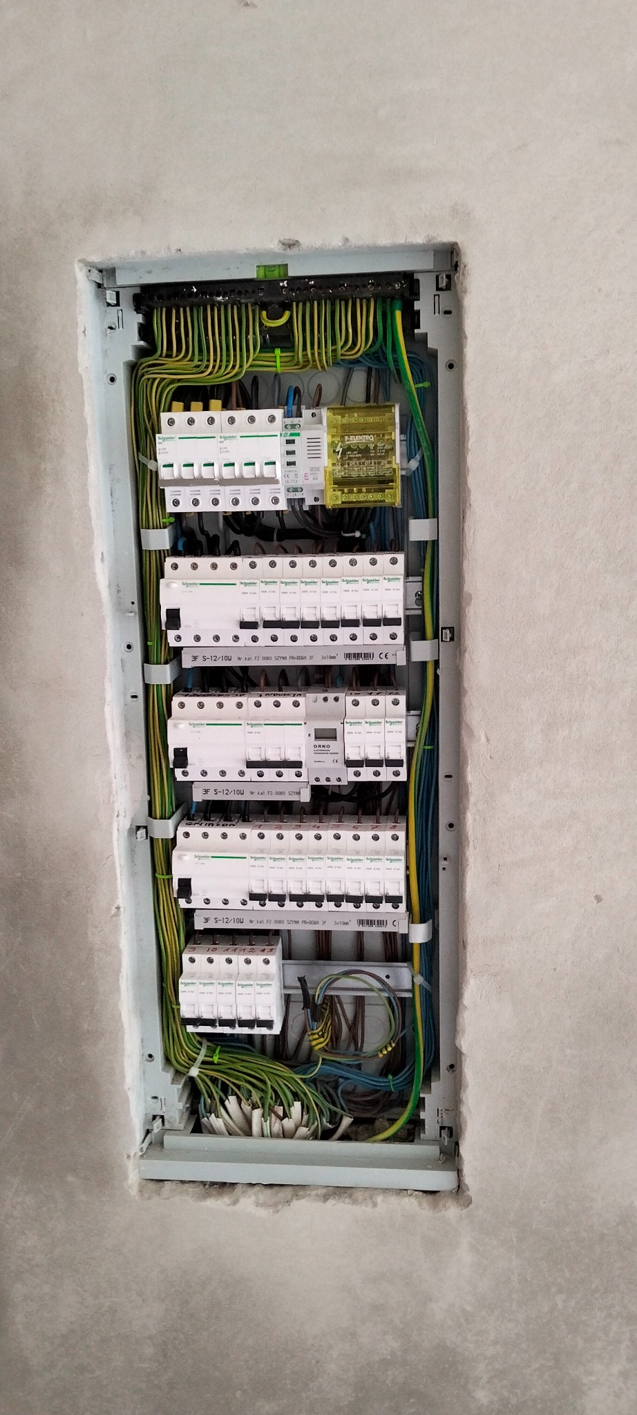 Rozdzielnica elektryczna z zamontowanymi bezpiecznikami i okablowaniem, umieszczona w ścianie w trakcie wykończenia. Widoczne przewody w kolorach żółto-zielonym.