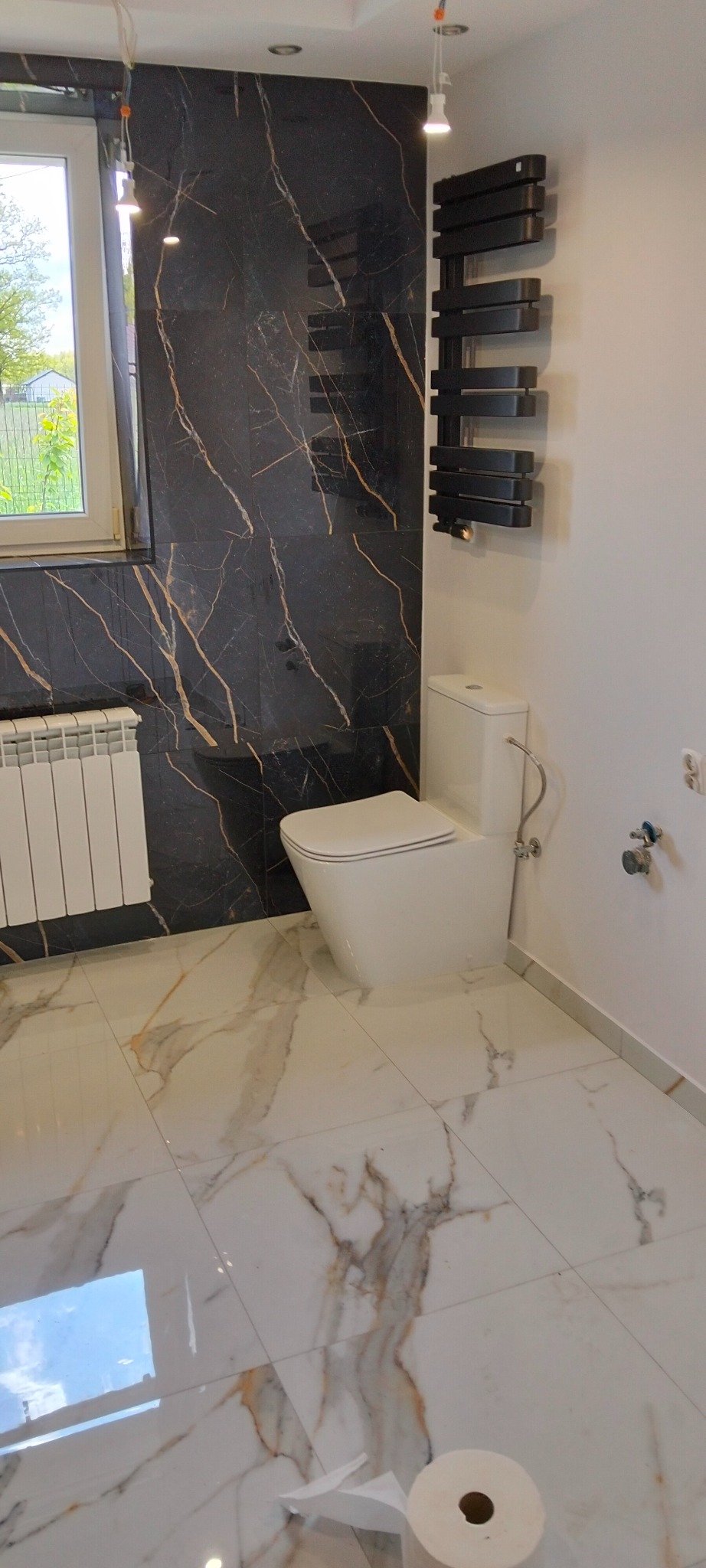 Wykończona łazienka z czarnymi płytkami imitującymi marmur z złotymi żyłami, białą armaturą i grzejnikiem drabinkowym. Błyszcząca podłoga z wzorem marmuru.
