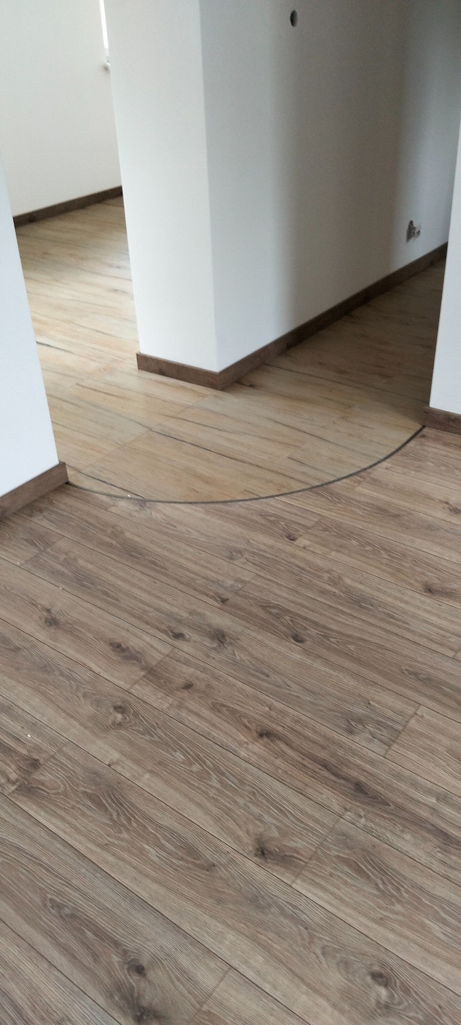 Ukończona podłoga z paneli w odcieniach brązu, z widocznymi listwami przypodłogowymi i fragmentem białej ściany. Widok na korytarz.