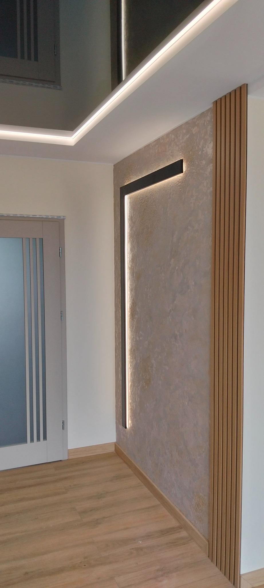 Wykończona ściana z dekoracyjnym tynkiem, oświetleniem LED w kształcie litery L i drewnianymi lamelami. Widoczna podłoga z paneli i fragment drzwi.