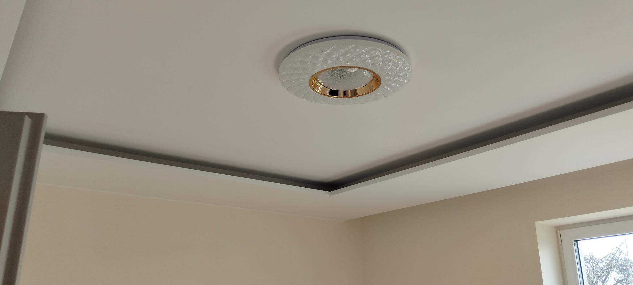 Nowoczesna lampa sufitowa z dekoracyjną obudową i złotym akcentem, wkomponowana w podwieszany sufit z szarą listwą. Jasne ściany i okno w tle.