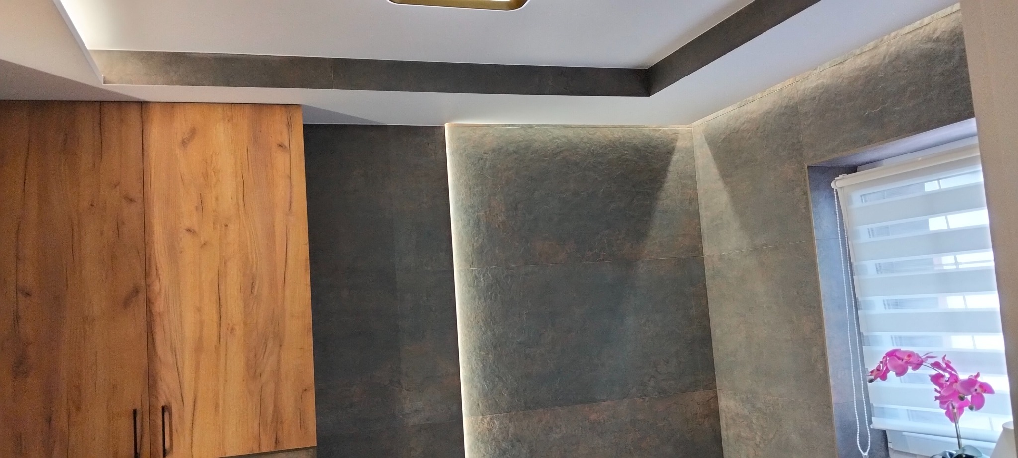 Nowoczesna łazienka z drewnianą szafką, szarą płytką imitującą kamień i subtelnym oświetleniem LED. W oknie roślina z różowymi kwiatami.