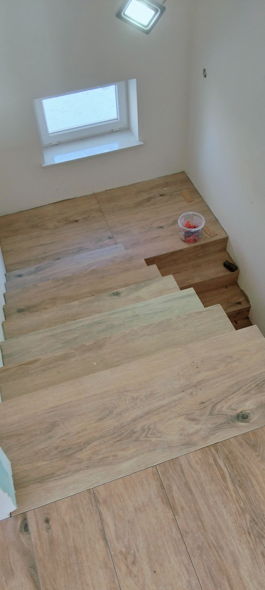 Wykończone schody z drewnopodobnych płytek, widok z góry. Na podeście wiaderko z krzyżykami dystansowymi i poziomica. Okno w tle. Jasne wnętrze.