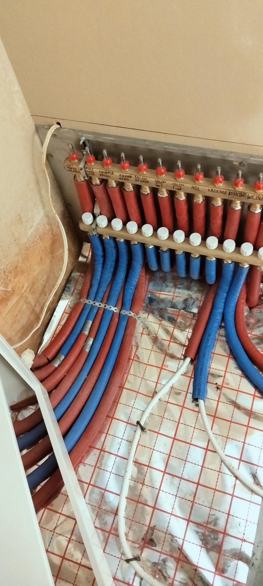 Rozdzielacz ogrzewania podłogowego z rurami w kolorze czerwonym i niebieskim, zamontowany na folii izolacyjnej z nadrukowaną siatką. Widoczne napisy na rozdzielaczu.