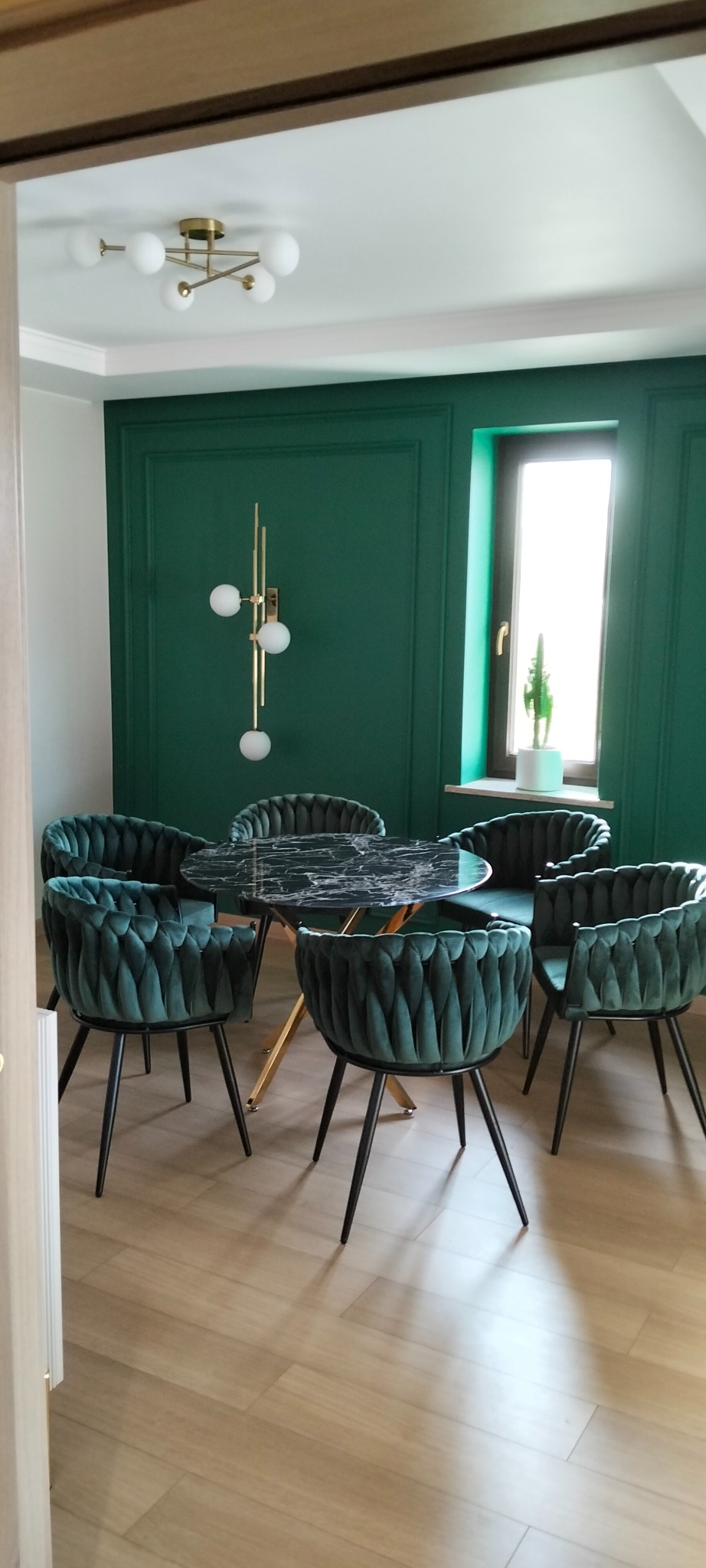 Szmaragdowe krzesła z weluru przy okrągłym stole z blatem imitującym marmur, na tle ściany w kolorze butelkowej zieleni z dekoracyjnymi panelami.