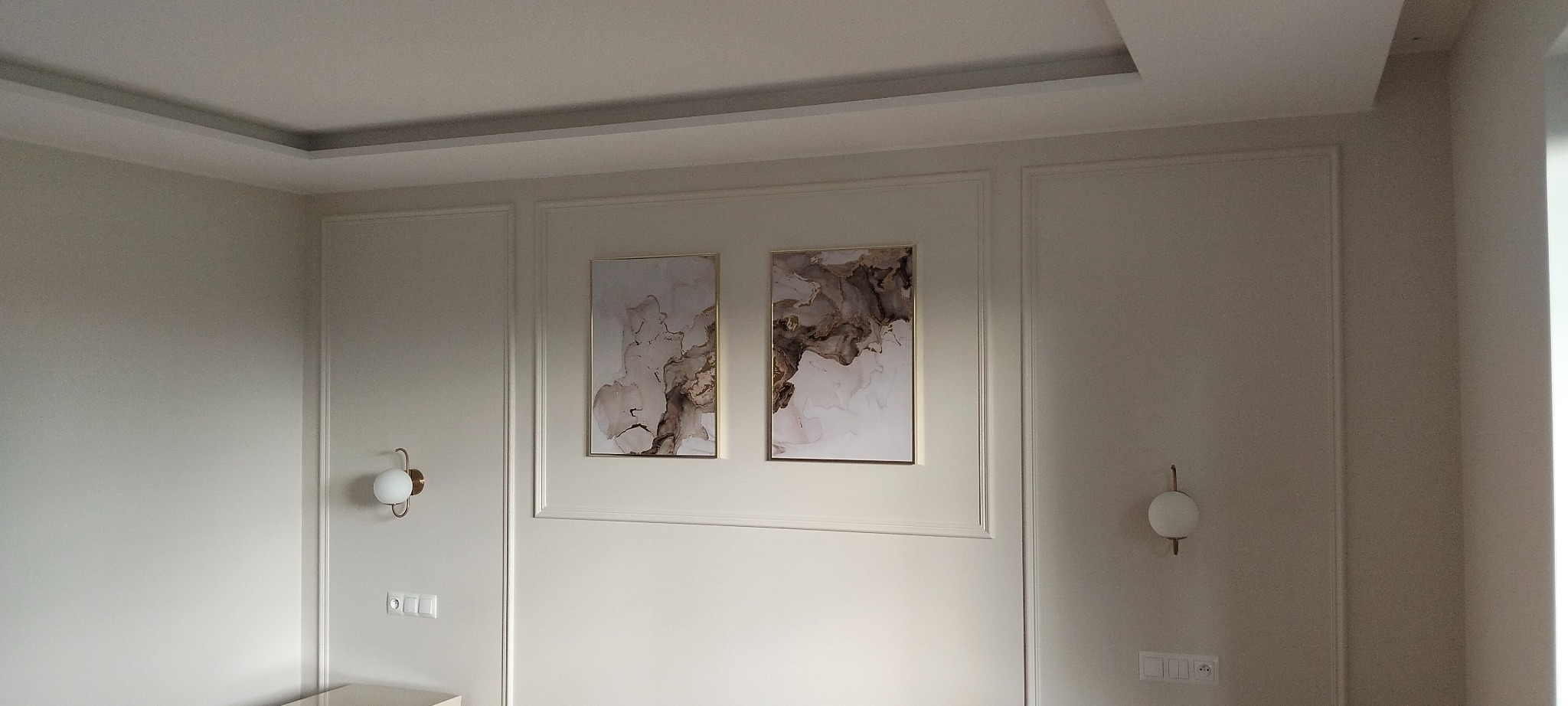 Eleganckie wykończenie ściany z dwoma abstrakcyjnymi obrazami w złotych ramach i symetrycznie rozmieszczonymi kinkietami w ciepłym odcieniu złota.