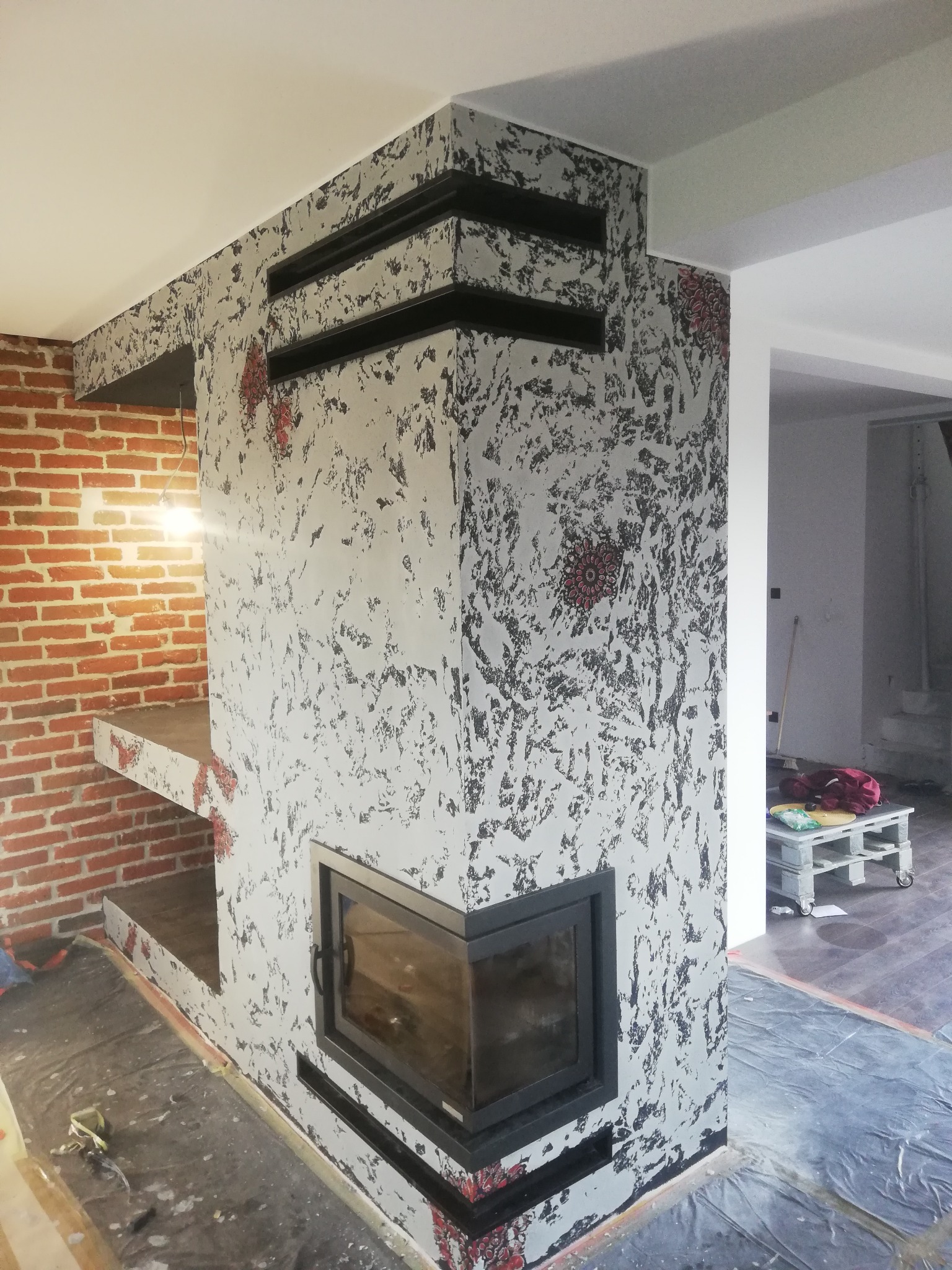 Wykończony kominek narożny z czarnym wkładem, obudowany dekoracyjnym tynkiem w odcieniach szarości i czerni, z elementami cegły. Widoczne zabezpieczenia podłogi.