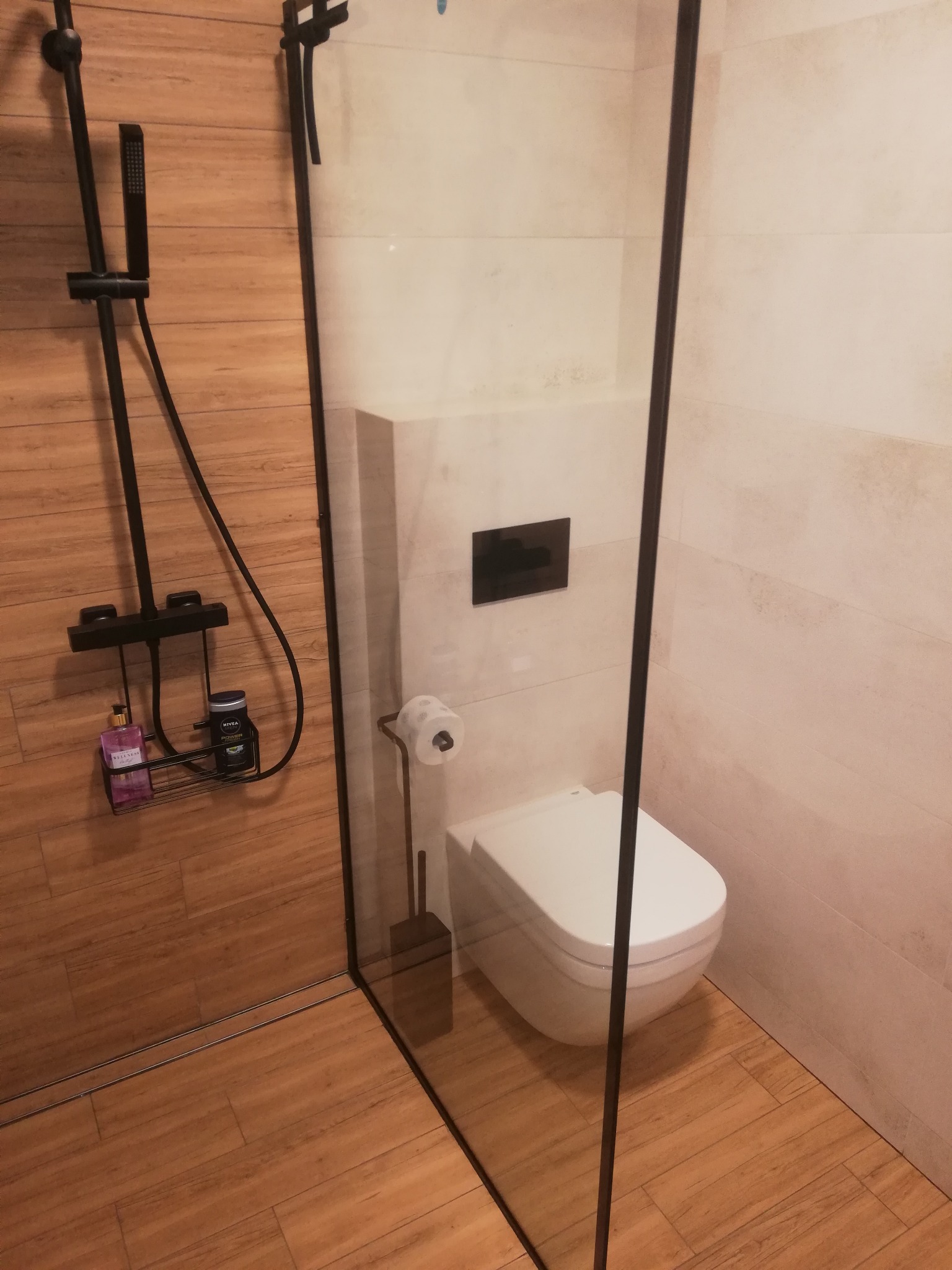 Nowoczesna kabina prysznicowa z czarną armaturą i drewnopodobnymi płytkami. Widoczna toaleta podwieszana i szklana ściana kabiny.