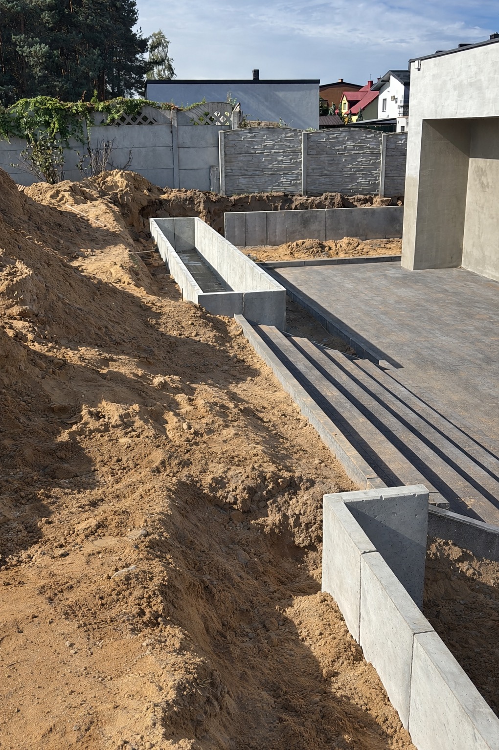 Przygotowanie terenu pod kostkę brukową z betonowymi obrzeżami. Wykop pod fundamenty ogrodzenia betonowego. Widoczne warstwy ziemi i częściowo ułożona kostka.