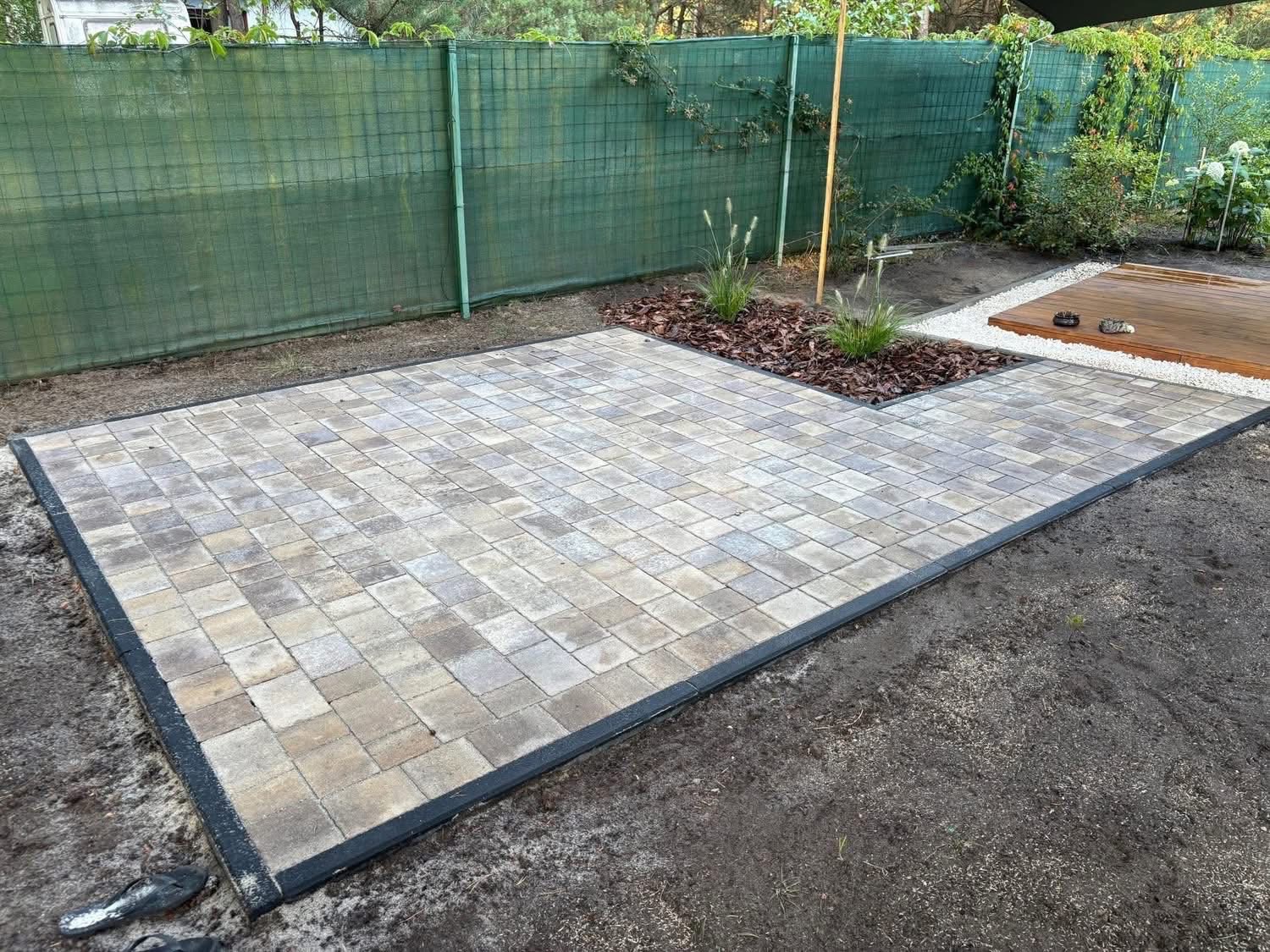 Ułożona kostka brukowa w ogrodzie, z czarną obwódką i rabatą z korą. Widoczny fragment drewnianego tarasu i zielone ogrodzenie siatkowe w tle.