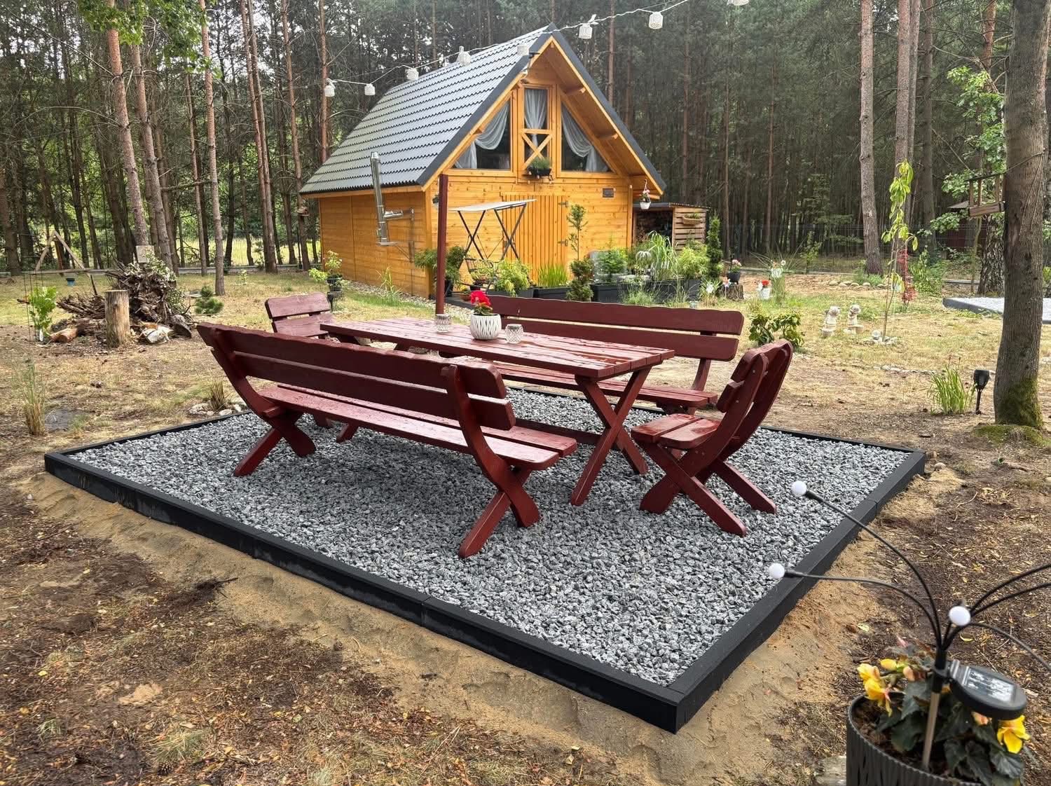 Ukończona strefa relaksu w ogrodzie: drewniany stół z ławkami na wysypanym kamieniem placu, obrzeża z czarnej kostki. W tle domek letniskowy i drzewa.