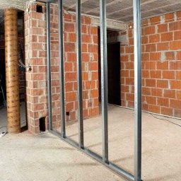 Wojciech Falenski - Montaż metalowej konstrukcji ścianki działowej na tle surowej cegły w remontowanym wnętrzu. Widoczne profile pionowe i poziome, surowy betonowy strop.