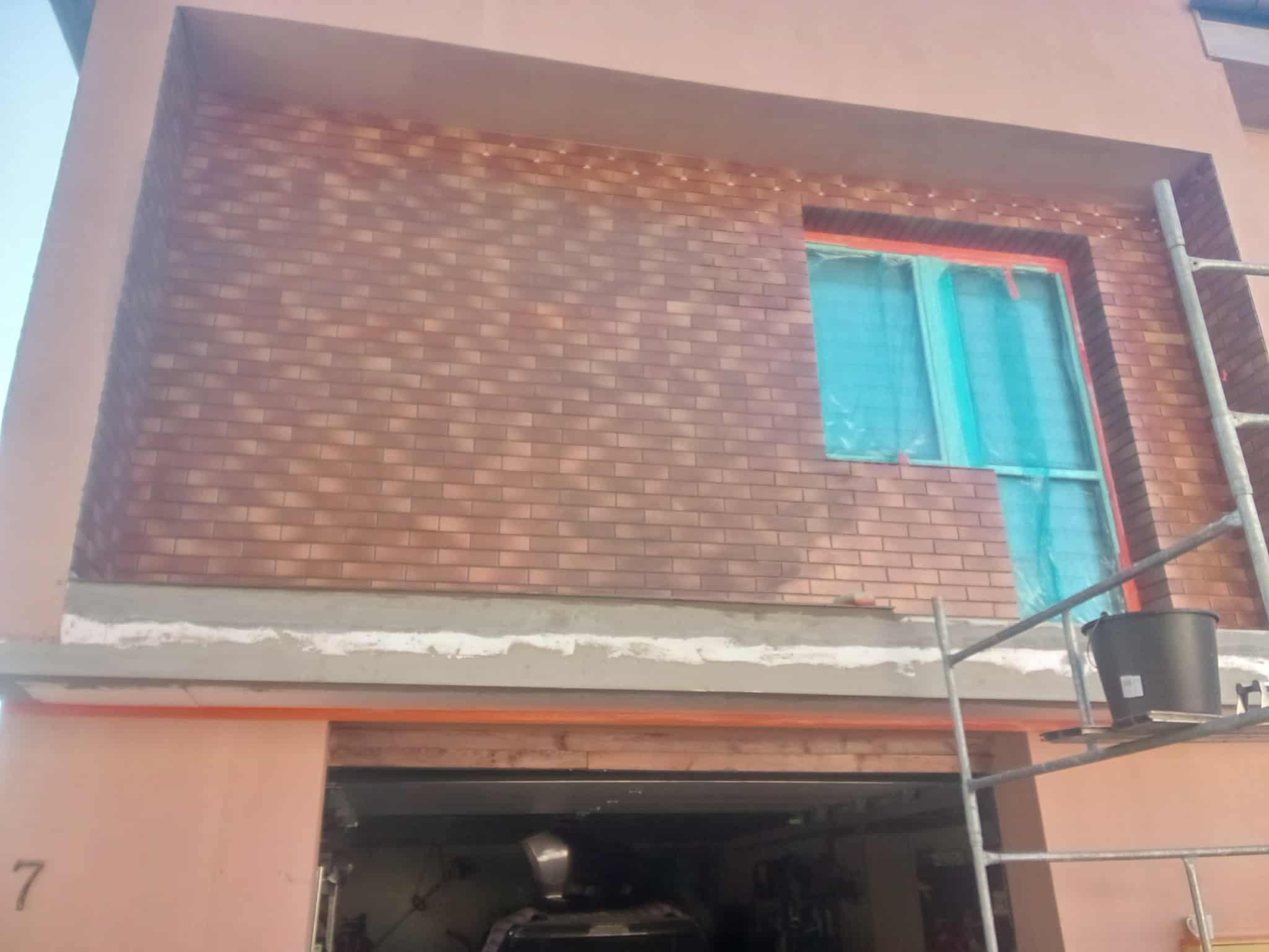 Elewacja budynku w trakcie wykańczania z cegły klinkierowej, okno zabezpieczone folią, widoczne rusztowanie i wiadro. Prace wykończeniowe na zewnątrz budynku.