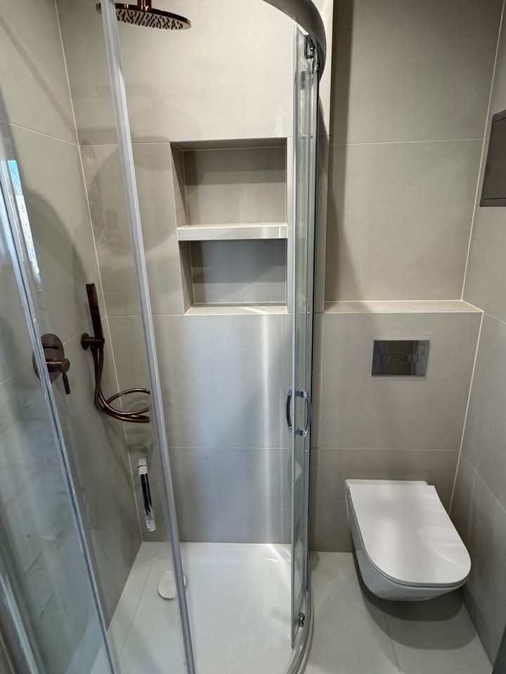 Nowoczesna łazienka z kabiną prysznicową typu walk-in, w odcieniach szarości. Widoczna toaleta podwieszana i wnęka z półkami w ścianie. Armatura w kolorze miedzi.