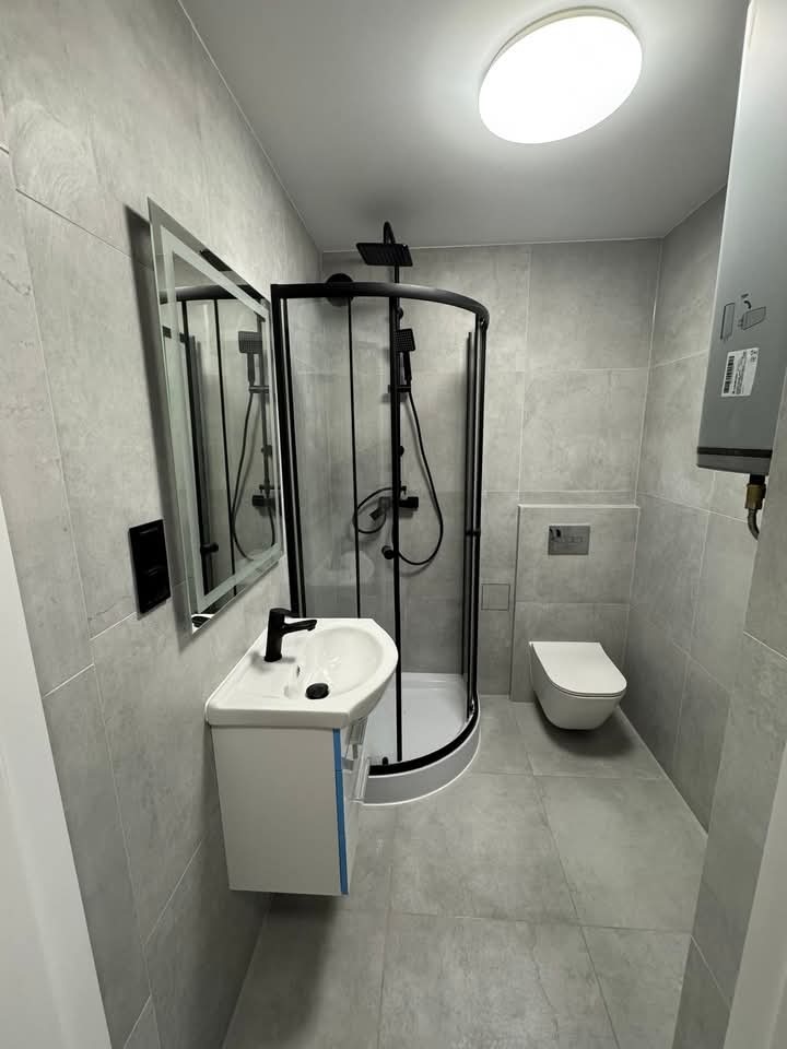 Nowoczesna łazienka w szarościach: kabina prysznicowa z czarnymi profilami, podwieszana umywalka z szafką i lustrem, toaleta. Minimalistyczny design, oświetlenie sufitowe.