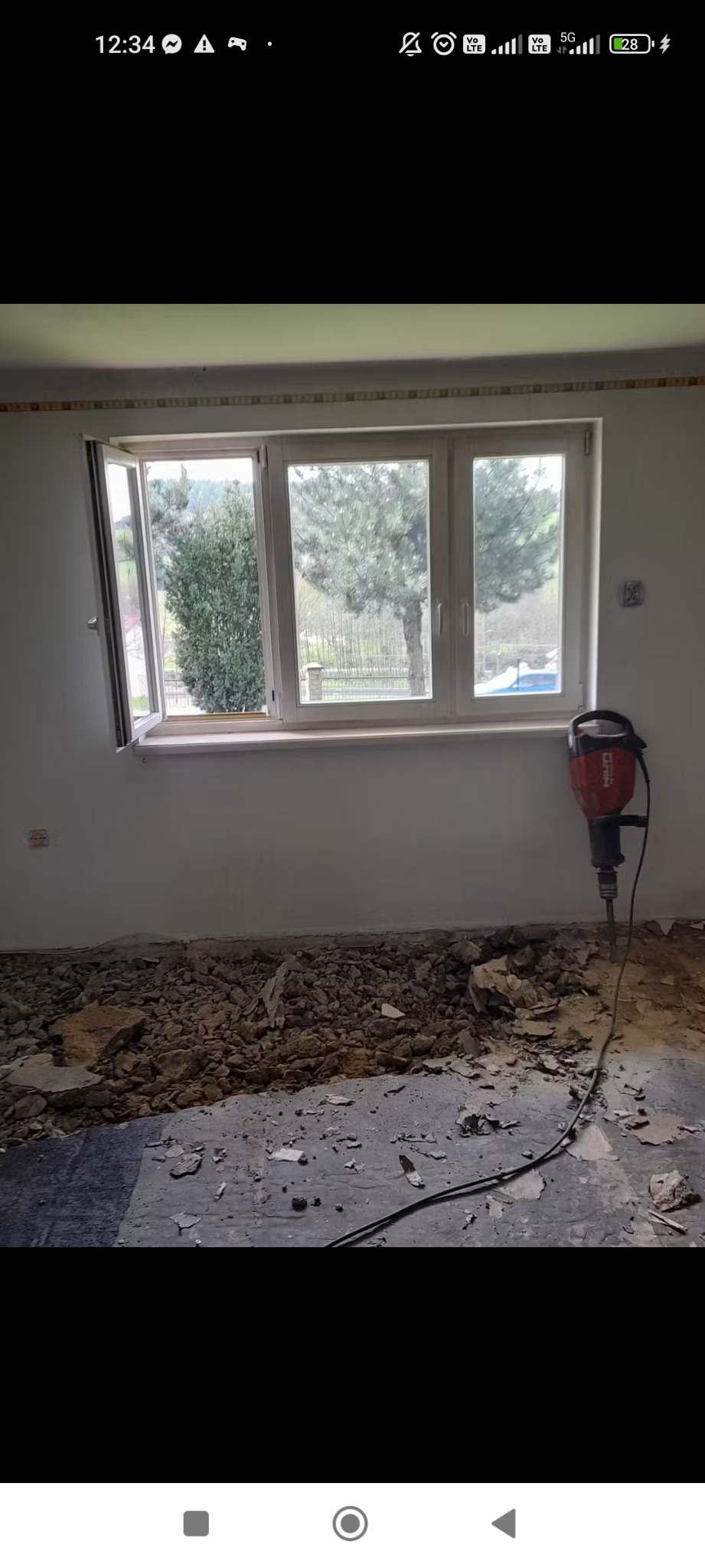 Demontaż podłogi w pokoju z oknem. Widoczny gruz i młot udarowy Hilti. Otwarta okiennica. Etap przygotowawczy do remontu.