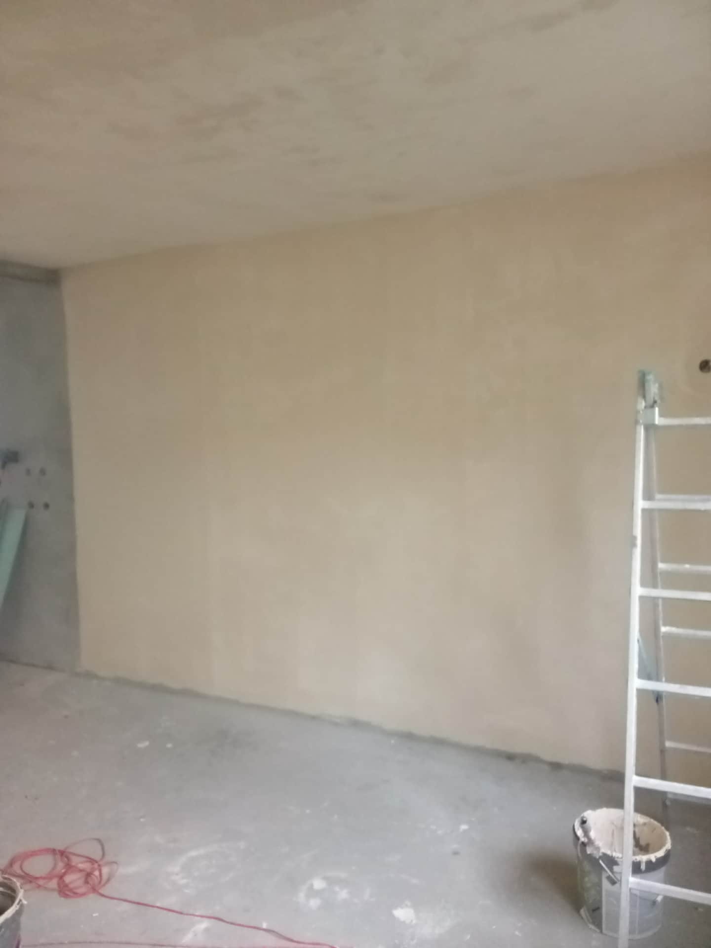 Ściana po nałożeniu gładzi, widoczna drabina malarska i wiadro z gładzią. Świeżo wygładzona powierzchnia gotowa do dalszych prac wykończeniowych.