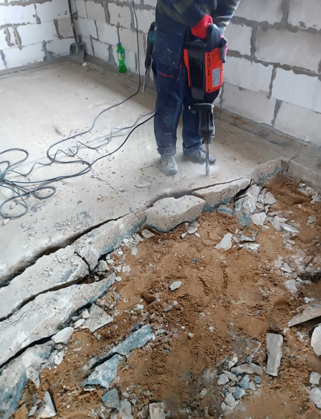 Demontaż posadzki w Oświęcimiu. Robotnik w kombinezonie używa młota pneumatycznego do skuwania betonowej podłogi. Widoczne gruz i odsłonięta ziemia.