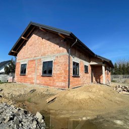 FHU GALAN Grzegorz Łączak - Surowy budynek z czarnymi oknami PCV w trakcie budowy, widoczne materiały budowlane i ziemia. Ujęcie w słoneczny dzień, błękitne niebo.