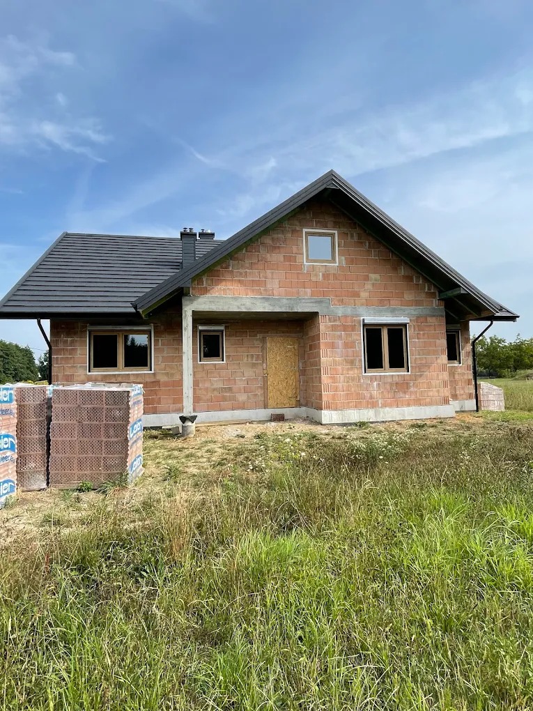 Surowy stan domu jednorodzinnego z zamontowanymi oknami w brązowych ramach. Na pierwszym planie palety z cegłami. Widok na budynek z zewnątrz, w tle zielona trawa i błękitne niebo.