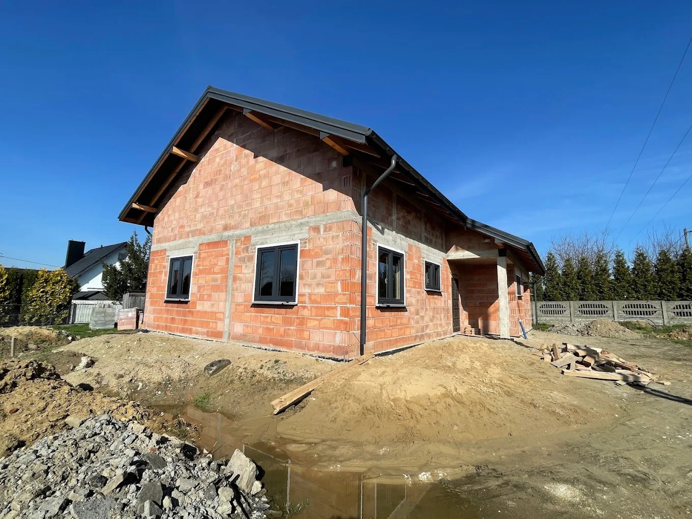 Surowy budynek z czarnymi oknami PCV w trakcie budowy, widoczne materiały budowlane i ziemia. Ujęcie w słoneczny dzień, błękitne niebo.