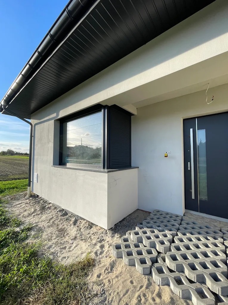 Nowoczesny dom jednorodzinny z czarną stolarką okienną i drzwiową. Widoczne okno z żaluzjami zewnętrznymi i drzwi wejściowe. Ułożona kostka ażurowa.