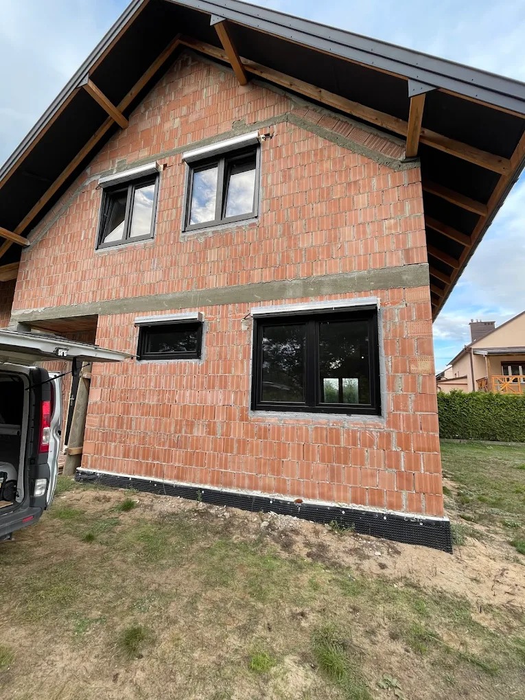 Surowy mur budynku z czarnymi oknami i częściowo opuszczonymi roletami zewnętrznymi. Widoczny fragment samochodu dostawczego. Dom w trakcie budowy.