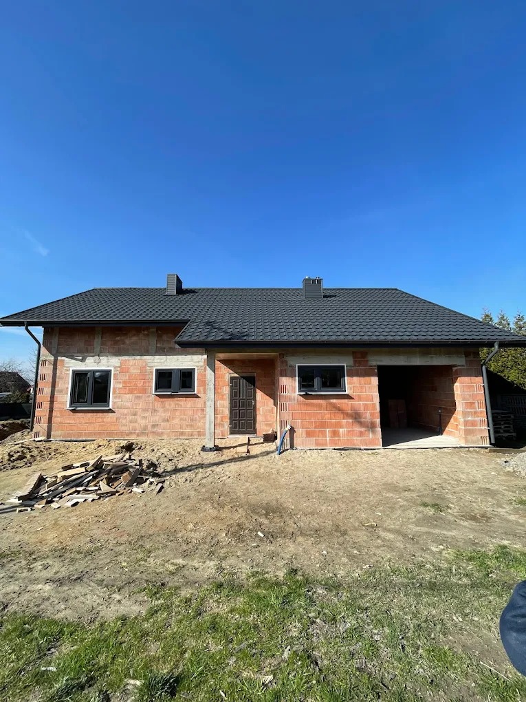 Surowy stan domu jednorodzinnego z czerwonej cegły, z zamontowanymi oknami i drzwiami, pod czarnym dachem, w słoneczny dzień. Widoczne materiały budowlane.