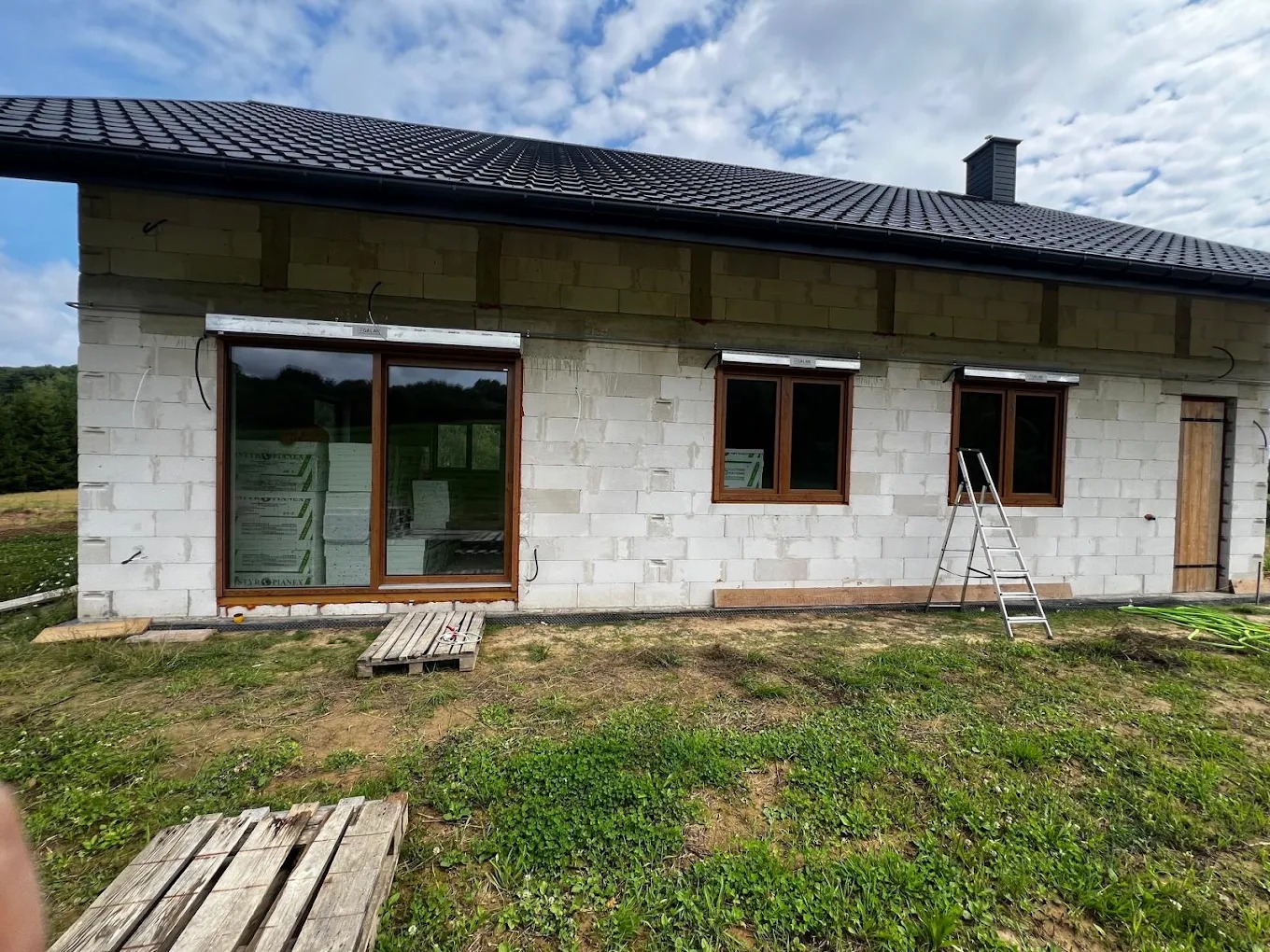 Dom w trakcie budowy z zamontowanymi oknami z brązowymi ramami i roletami zewnętrznymi. Widoczna drabina oparta o ścianę i palety na trawie.