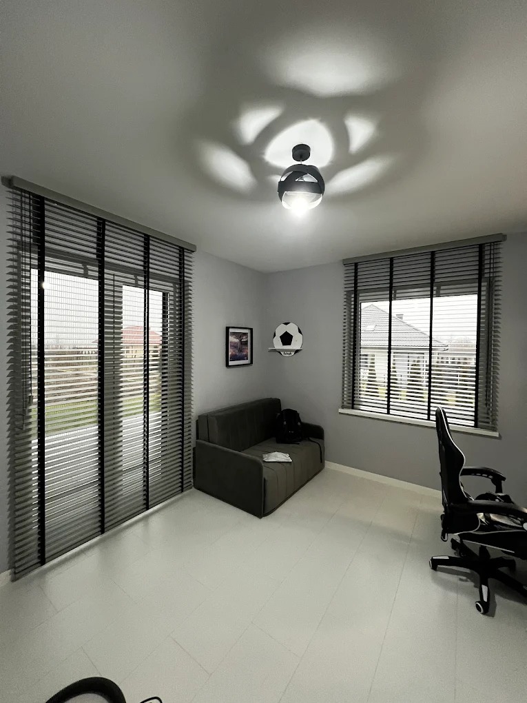 Pokój z szarymi żaluzjami drewnianymi na oknach i drzwiach balkonowych. Nowoczesna lampa sufitowa rzuca wzór na sufit. Minimalistyczny wystrój.