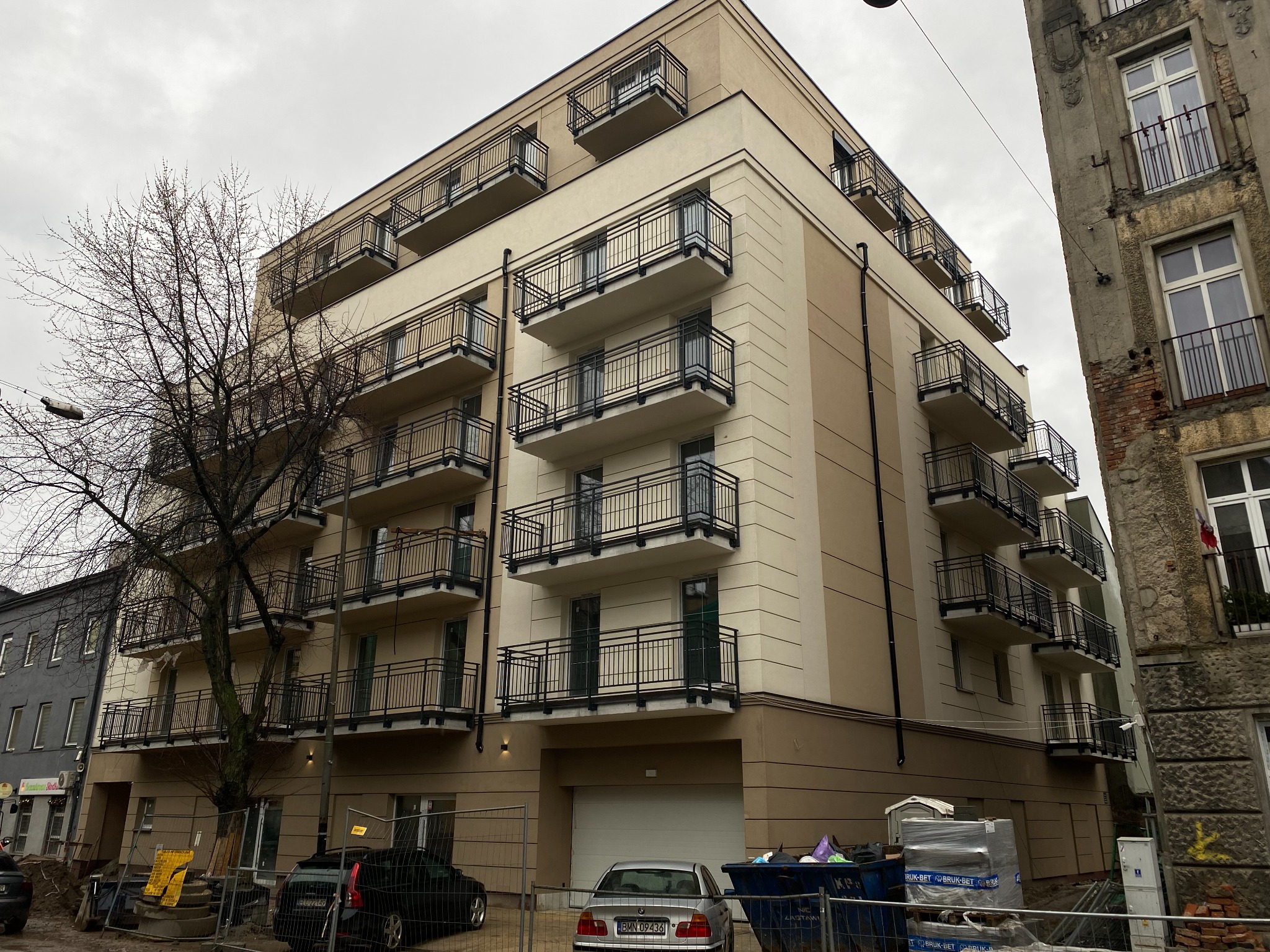 Nowoczesny budynek wielorodzinny z balkonami, w trakcie prac wykończeniowych. Widoczne ogrodzenie budowy, samochody i materiały budowlane. Pochmurne niebo w tle.