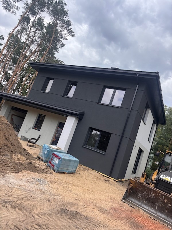 Nowoczesny dom z czarną elewacją i dużymi oknami, w trakcie prac wykończeniowych. Na pierwszym planie palety z materiałami budowlanymi i maszyna budowlana.