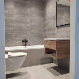 A&M System - Nowoczesna łazienka z szarymi płytkami, wiszącą szafką z umywalką i toaletą. Minimalistyczny design, czarne akcenty armatury, lustro z oświetleniem.