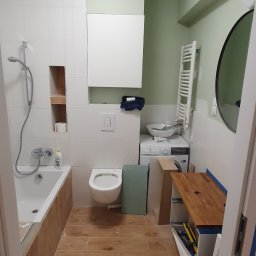 A&M System - Remont łazienki w trakcie: białe płytki, drewnopodobna podłoga, częściowo zamontowana armatura, widoczne narzędzia i materiały budowlane. Ściany w odcieniach zieleni.