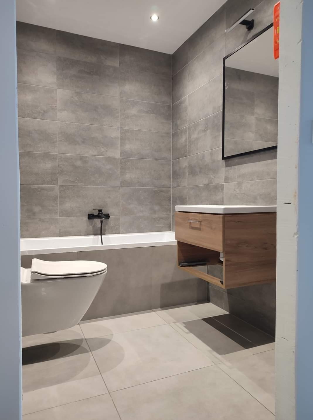 Nowoczesna łazienka z szarymi płytkami, wiszącą szafką z umywalką i toaletą. Minimalistyczny design, czarne akcenty armatury, lustro z oświetleniem.