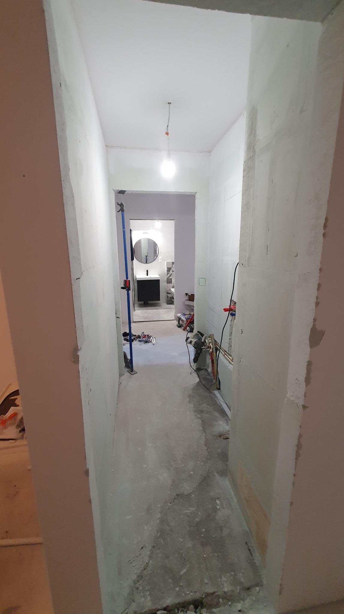 Korytarz w trakcie remontu z widoczną łazienką w tle. Ściany w stanie surowym, widoczne instalacje i narzędzia. Oświetlenie sufitowe.