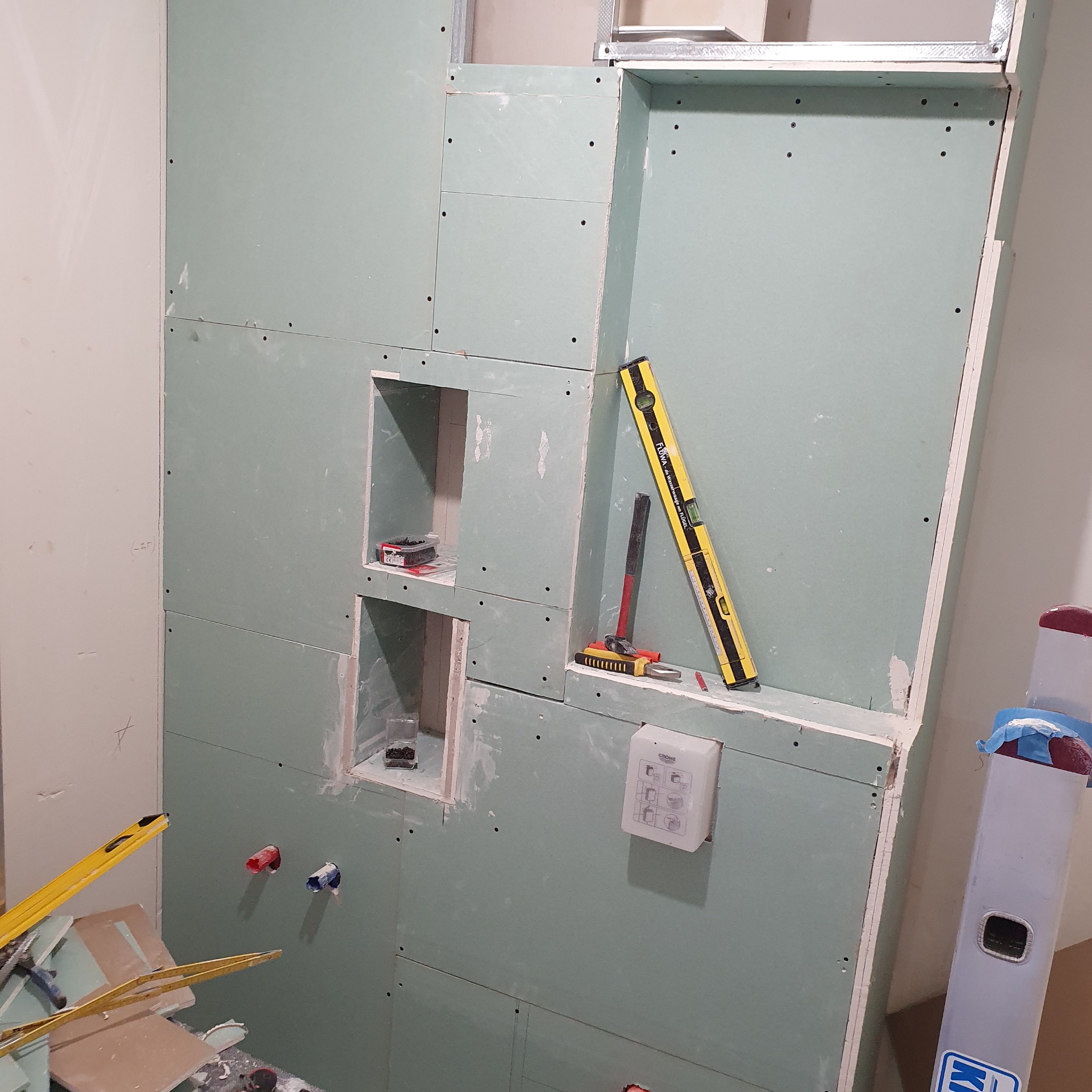Ściana z płyt karton-gips z wnękami, widoczne narzędzia (poziomica, młotek), instalacja wodna. Etap prac wykończeniowych w łazience.