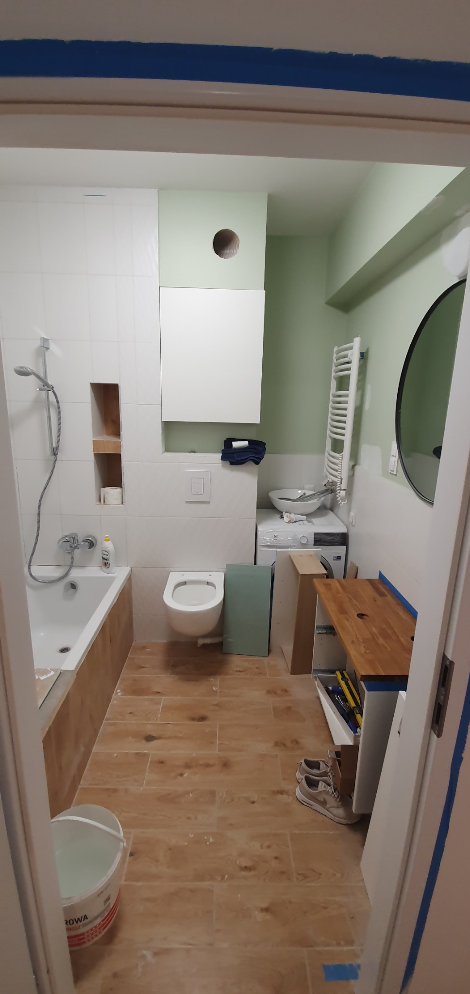 Remont łazienki w trakcie: białe płytki, drewnopodobna podłoga, częściowo zamontowana armatura, widoczne narzędzia i materiały budowlane. Ściany w odcieniach zieleni.