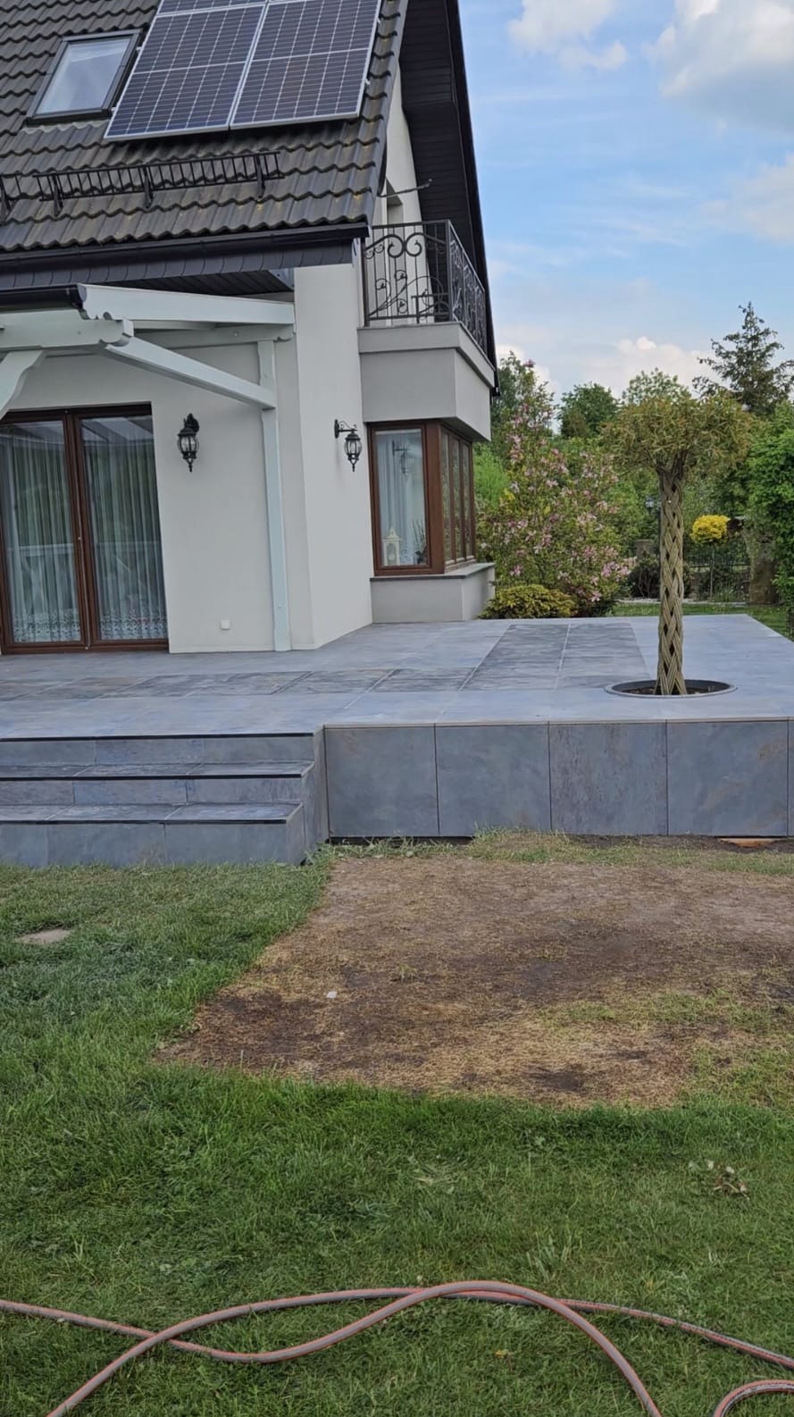 Nowoczesny taras z szarych płyt, zintegrowany z bryłą domu. Widoczne schody, drzewo wkomponowane w taras i fragment elewacji z oknami. Na dachu panele słoneczne.